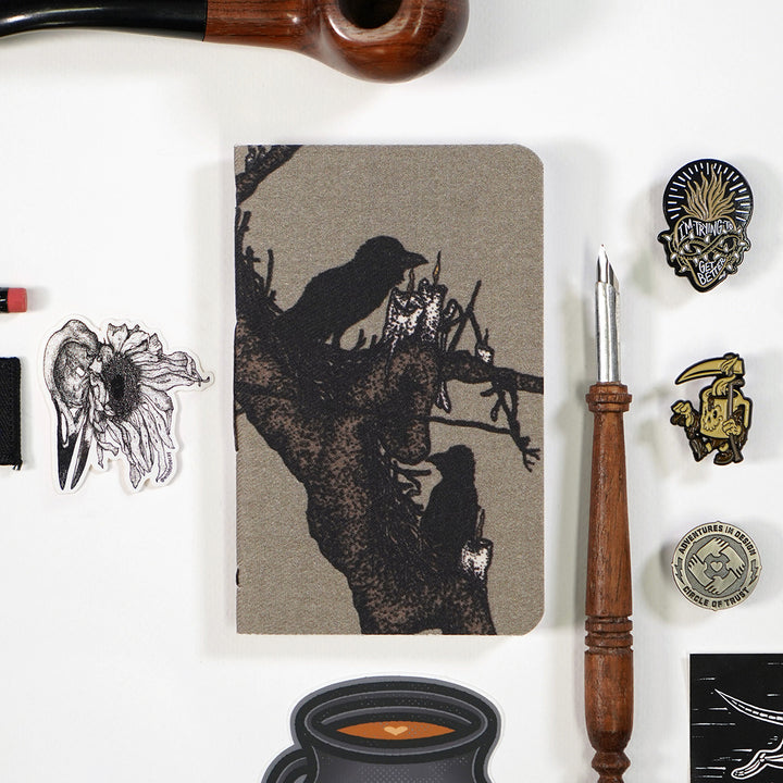 notebook cover with beige background and an illustration of two crows sitting on fat tree branches that also have candles on them. the notebook is surrounded by various objects line pins, stickers, and a tobacco pipe