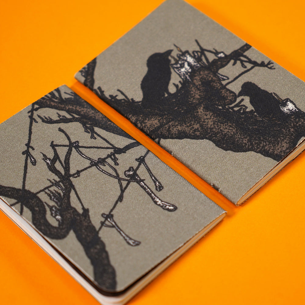 pocket notebook showing the design on both front and back covers. with birds in a tree, candles resting on the branches, and bones hanging from strings stretched between the branches