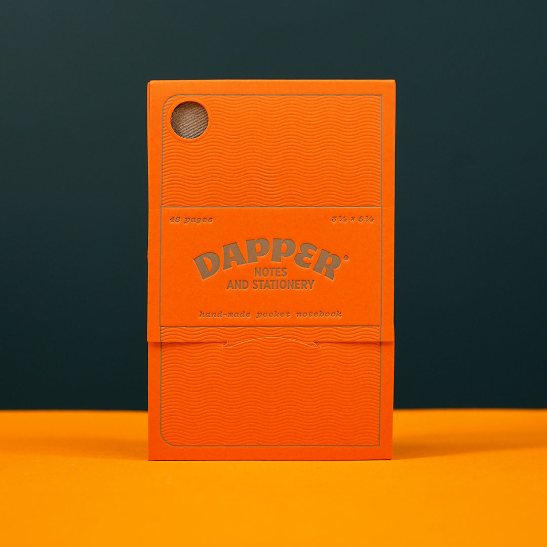 orange paper packaging for a pocket notebook standing on a yellow tabletop. the packaging reads 'dapper notes and stationery' and is printed with letterpressed textures by mamas sauce