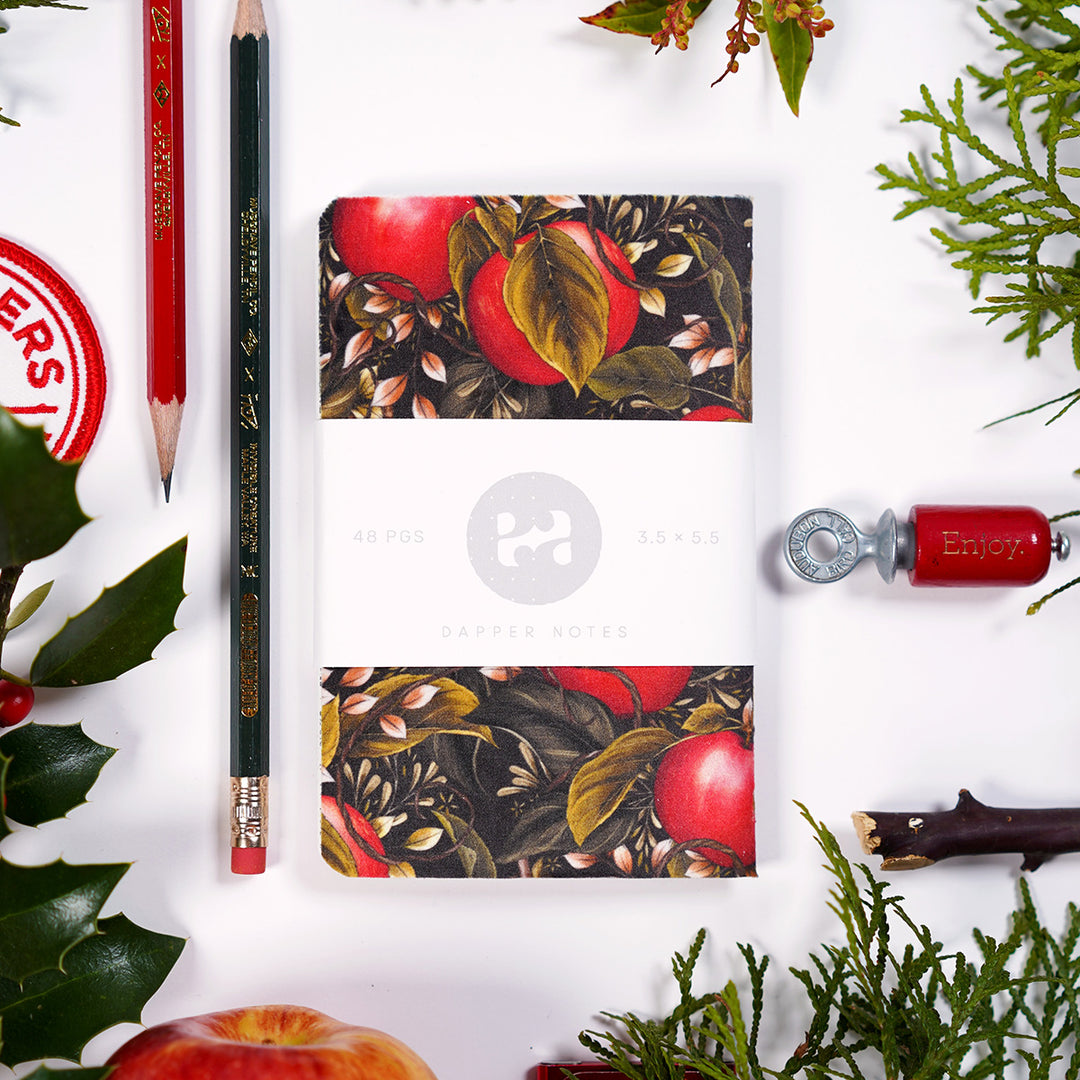 Pocket notebook with an apple and foliage themed cover, presented on a tabletop alongside other items like musgrave pencils, a bird call, and chirstmas-ey plants